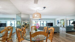 Un salon moderne avec un coin repas, comprenant une table ronde en verre, des chaises en bois, un luminaire suspendu en fibres tressées et un grand miroir qui reflète l’espace.