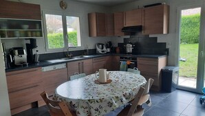A modern kitchen with wooden cabinets, a round table with floral tablecloth, and a view of the garden through the window. - Large family home in Annecy 6 bedrooms 12 people 15 minutes from the lake! (Annecy)