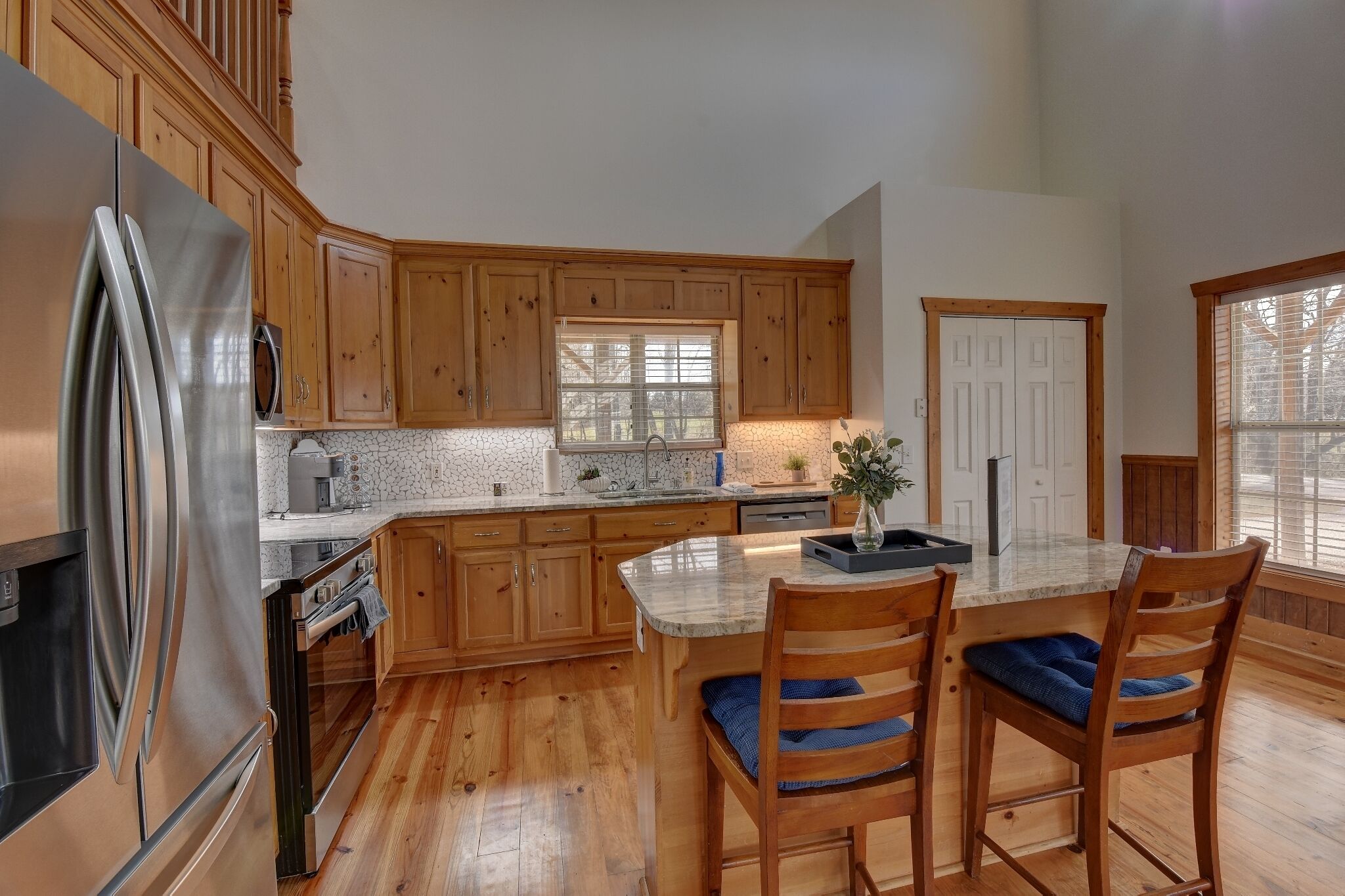 A kitchen with wooden cabinets, a stainless steel refrigerator, a marble countertop, and a dining area with two chairs.