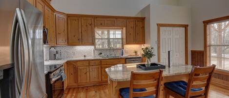 A kitchen with wooden cabinets, a stainless steel refrigerator, a marble countertop, and a dining area with two chairs.