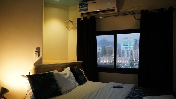 Ein Hotelzimmer mit Bett, Nachttisch, Lampe, Klimaanlage und Fenster mit Stadtblick.