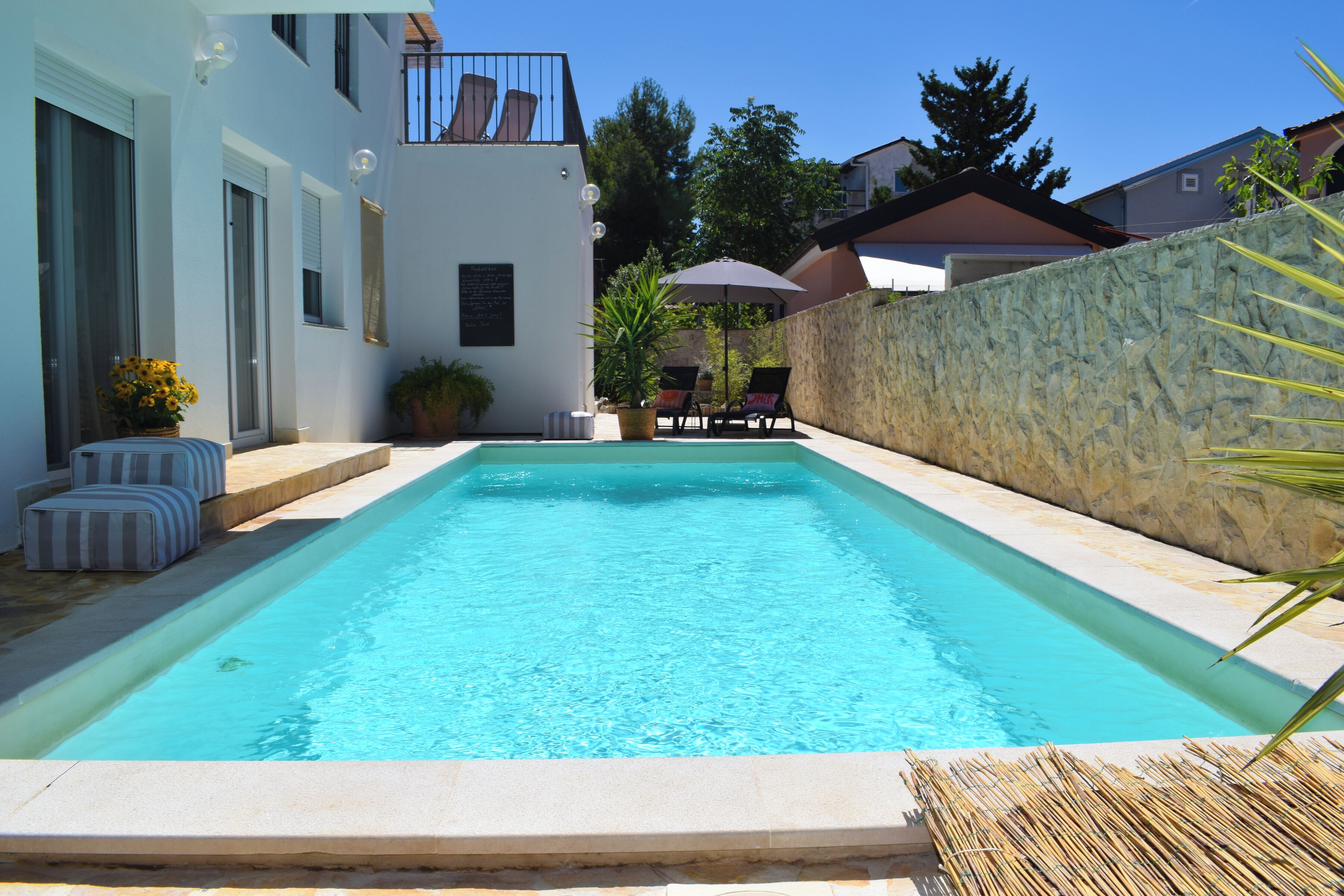 A rectangular swimming pool with a stone wall on one side and a white building with a balcony on the other.