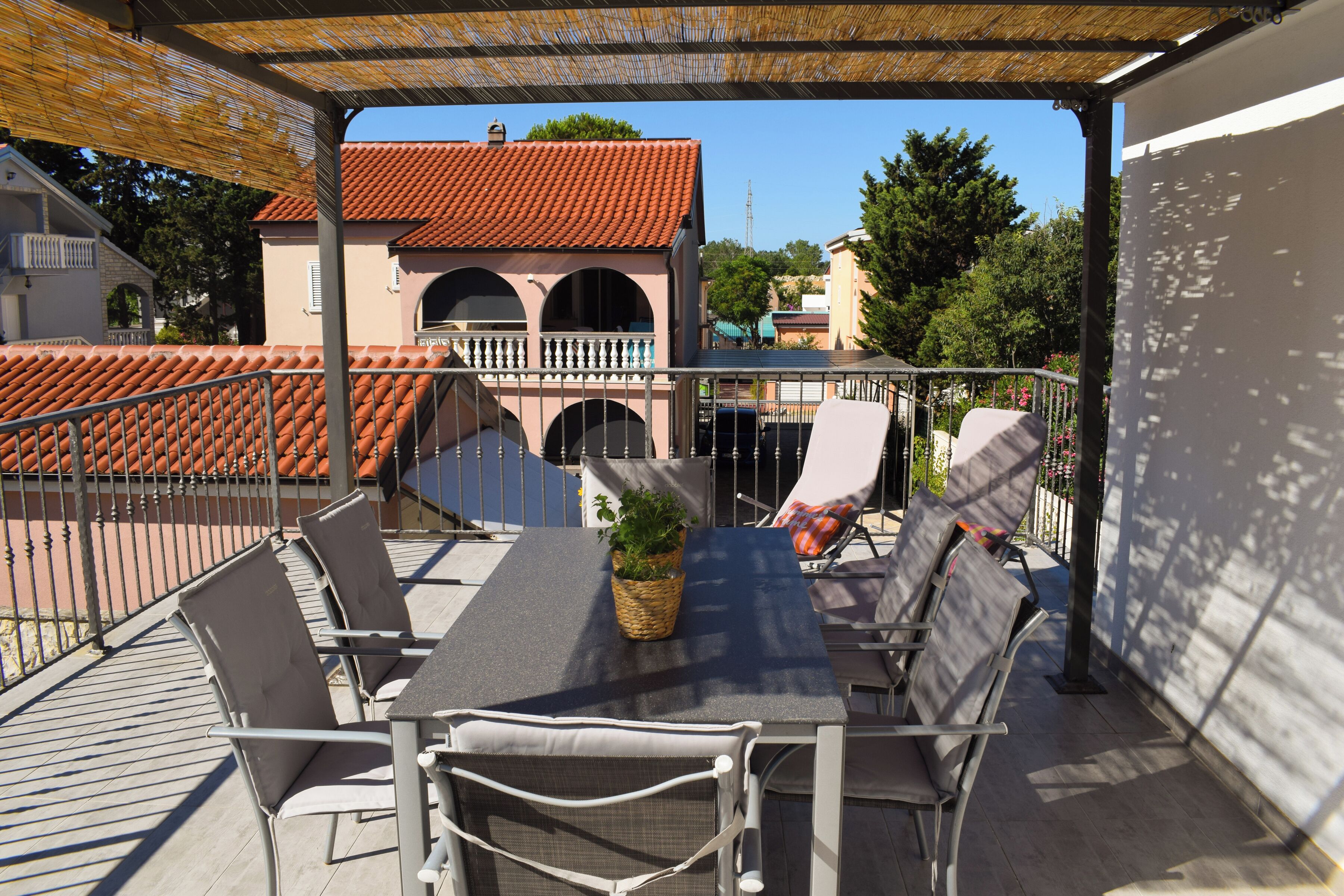 An outdoor patio with a table and chairs, a wicker basket with greenery, and a building with a red-tiled roof in the background.