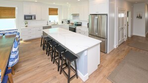 A modern kitchen with white cabinets, a large island with bar stools, stainless steel appliances, and a dining area with a blue and white tablecloth. - BIG VIEW. New 2026. 7 BR/7.5 BT, private 25'x15' pool, elevator, Surfside Beach (Surfside Beach)