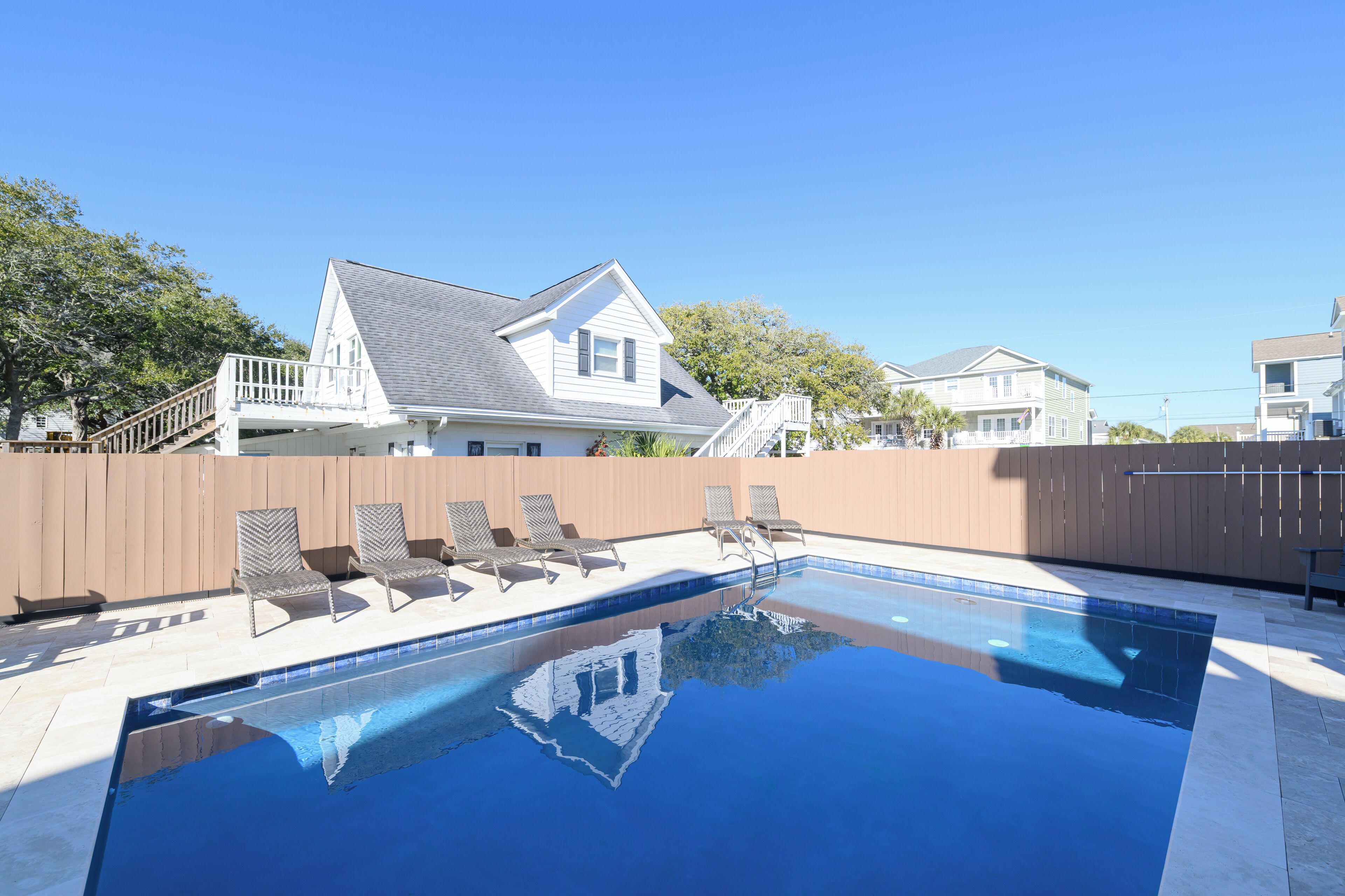 A swimming pool with lounge chairs and a wooden deck, surrounded by a wooden fence and houses in the background.