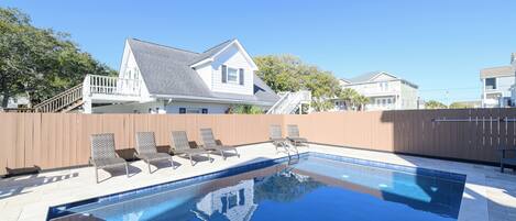 A swimming pool with lounge chairs and a wooden deck, surrounded by a wooden fence and houses in the background.