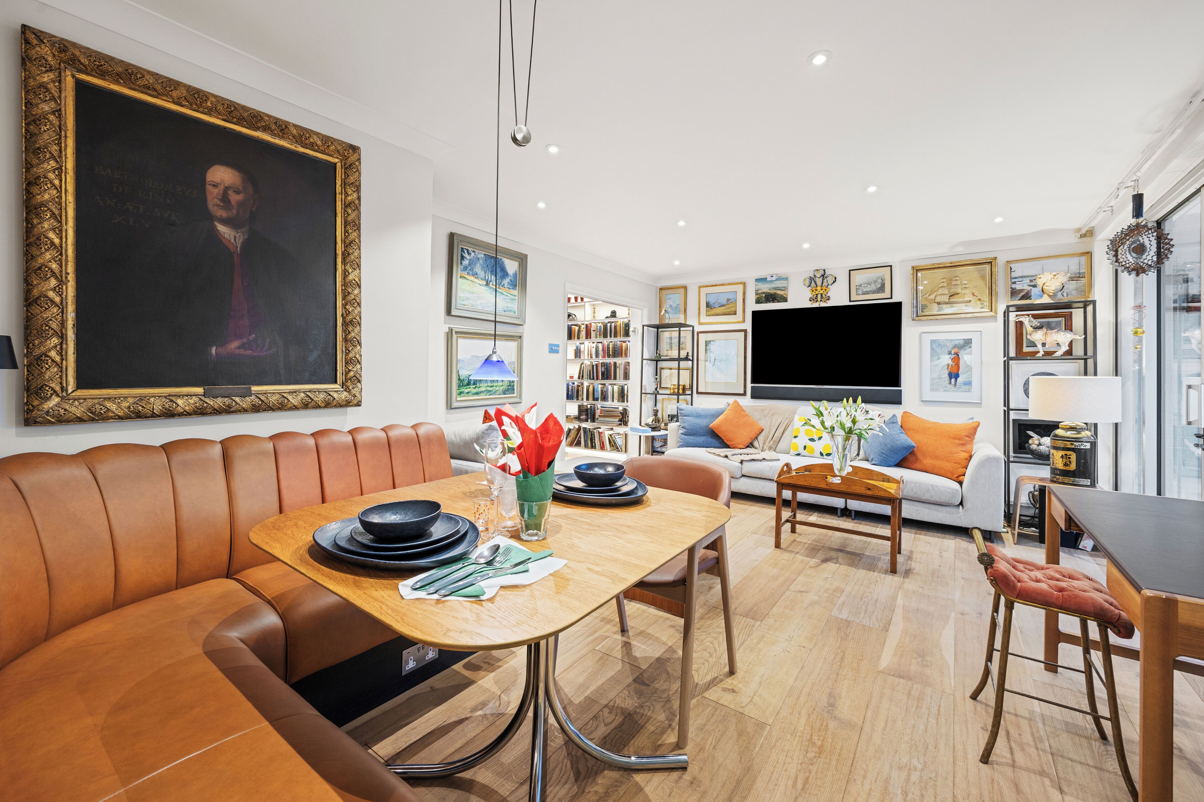 A modern living room with a dining area, featuring a large painting, bookshelves, and a flat-screen TV.