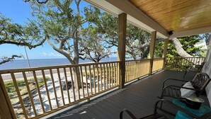 Een houten terras met een balustrade, met uitzicht op een strand en de oceaan. Er staan twee stoelen en een tafel met een blauw kussen.