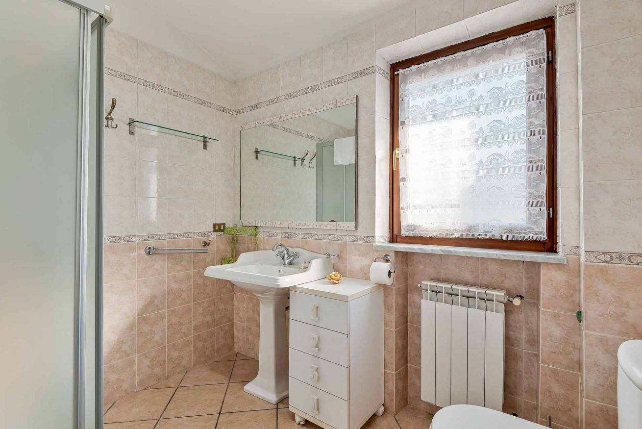 Ein Badezimmer mit Duschkabine aus Glas, einem Waschbecken auf Säule, einem Schrank mit Schubladen, einer Toilette und einem Heizkörper.