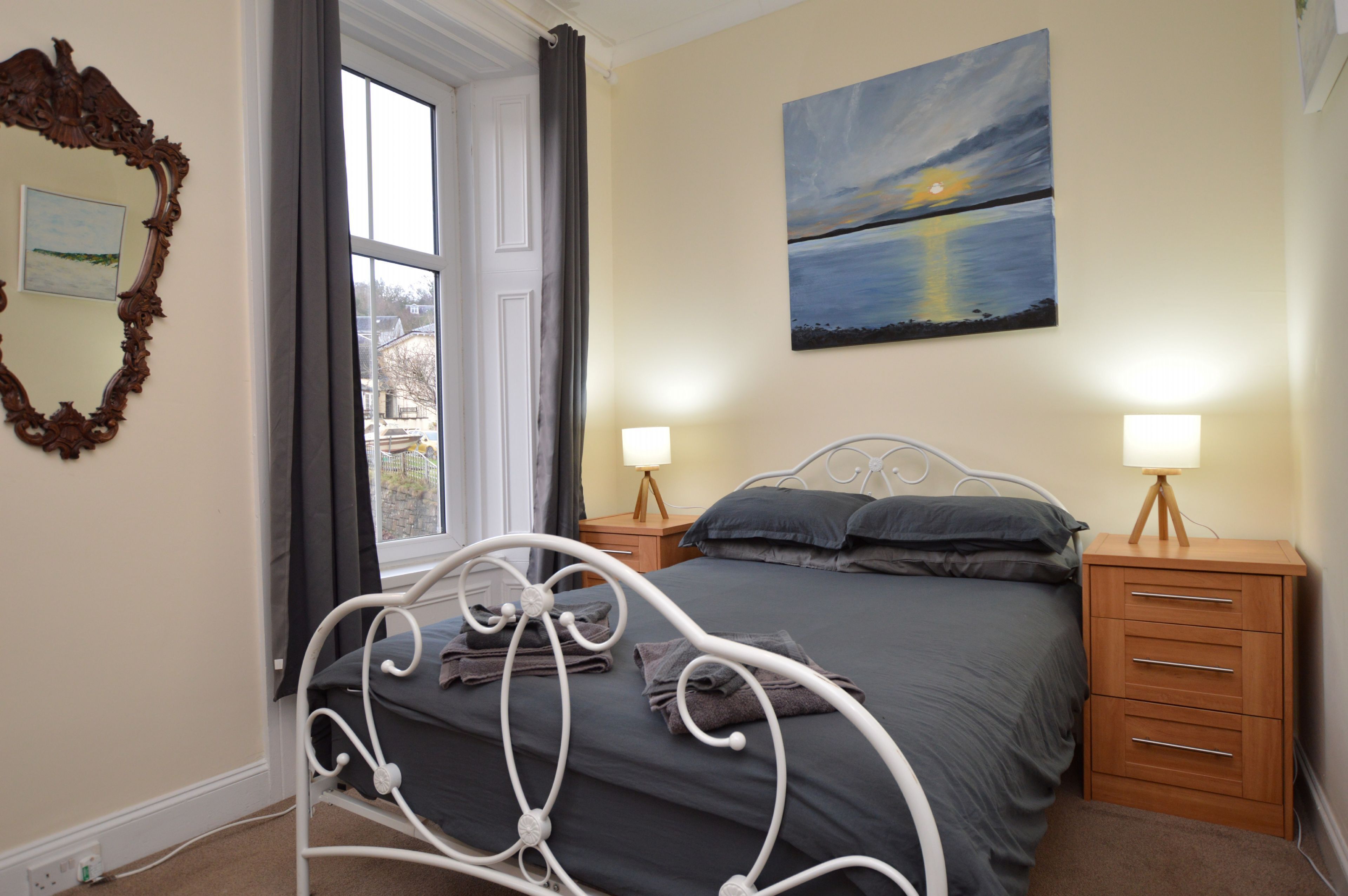 A bedroom with a bed, bedside lamps, a wooden nightstand, a mirror, and a painting of a sunset over water.