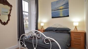 A bedroom with a bed, bedside lamps, a wooden nightstand, a mirror, and a painting of a sunset over water.