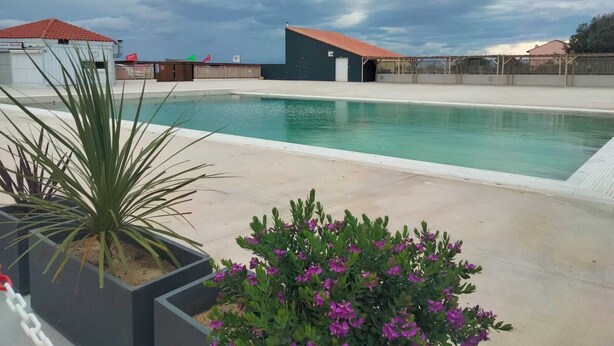 Une piscine dotée d’une terrasse carrelée, entourée d’une clôture, et un bâtiment avec un toit rouge en arrière-plan.