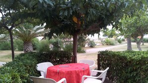 Une terrasse extérieure avec une table ronde recouverte d’une nappe rouge et quatre chaises en plastique blanches.