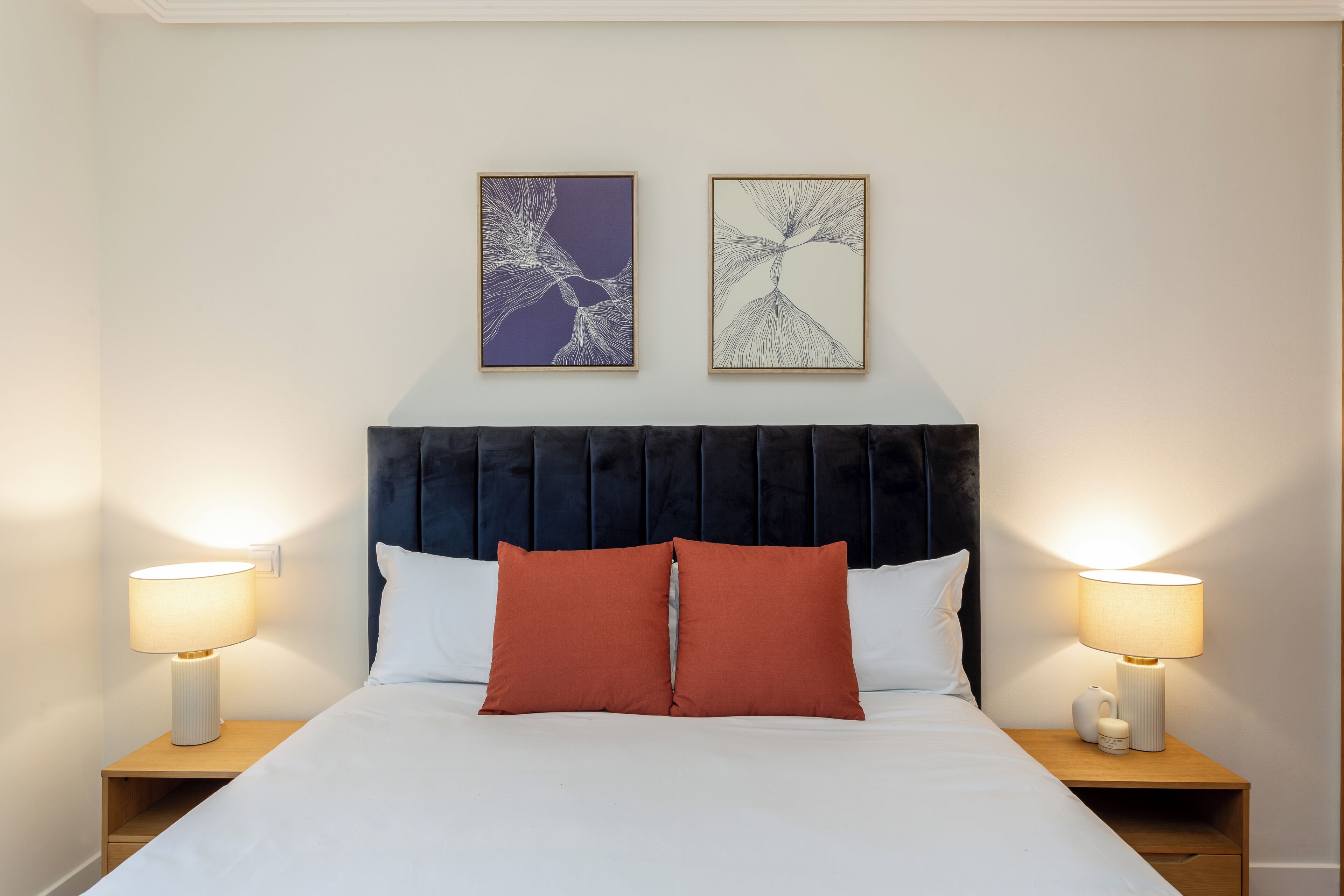 Una cama bien hecha con un cojín blanco y rojo, dos lámparas de noche y dos obras de arte enmarcadas en la pared.
