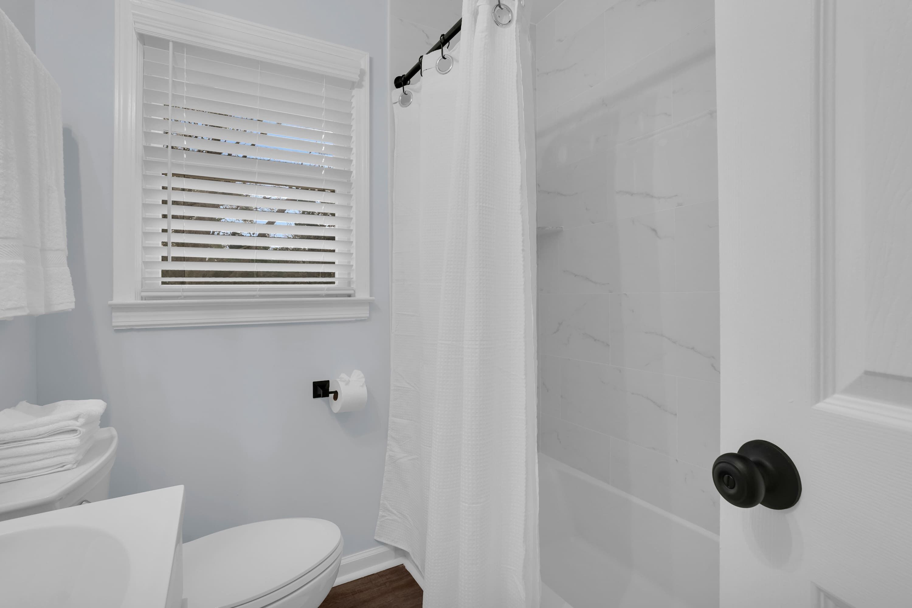 Une salle de bain avec des toilettes, une fenêtre avec des stores, un rideau de douche et une porte dotée d’une poignée ronde.