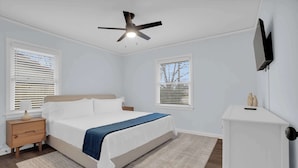 Ein Schlafzimmer mit Bett, Nachttisch, Kommode, Deckenventilator und zwei Fenstern mit Jalousien.