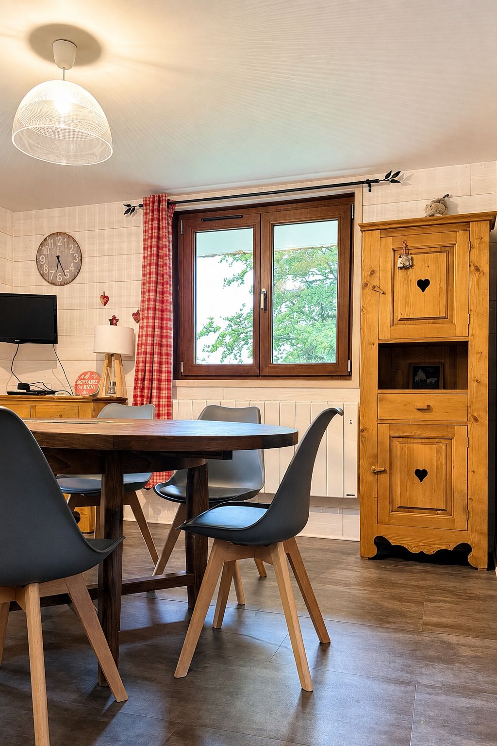 Un coin repas avec une table et des chaises en bois, un luminaire suspendu, une fenêtre offrant une vue agréable et un meuble en bois orné d’une découpe en forme de cœur.