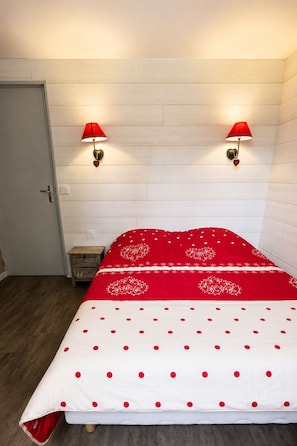 Un dormitori amb un llit, dues làmpades penjades a la paret, una tauleta de nit de fusta i una porta.