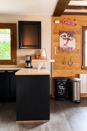 A kitchen with a black island, wooden cabinets, a coffee maker, and a poster on the wall. - Upstairs apartment, chalet on the slopes of Les Rousses (Prémanon)