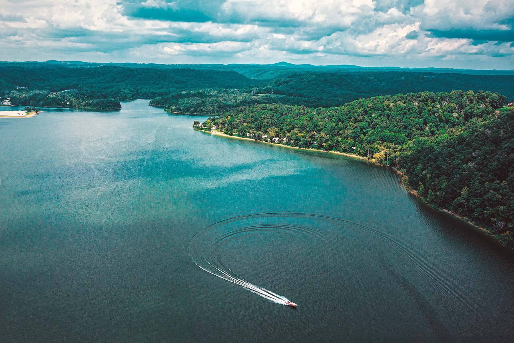 Une vue aérienne d’un lac entouré de collines boisées, avec un bateau qui laisse une vague derrière lui sur l’eau.