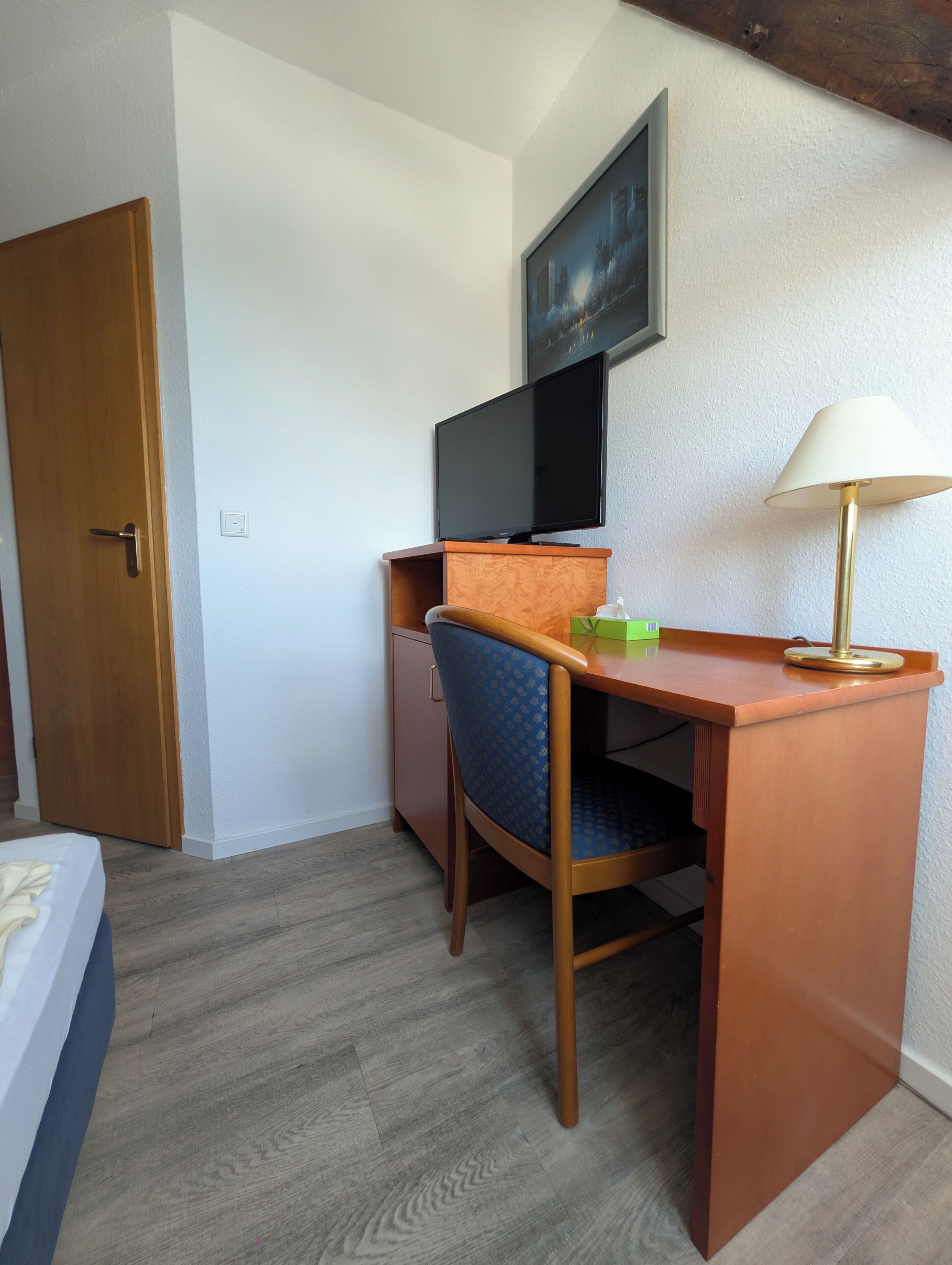 Une chambre d’hôtel de taille modeste, avec un bureau en bois, un téléviseur fixé au-dessus, une chaise, une table de chevet et une lampe.