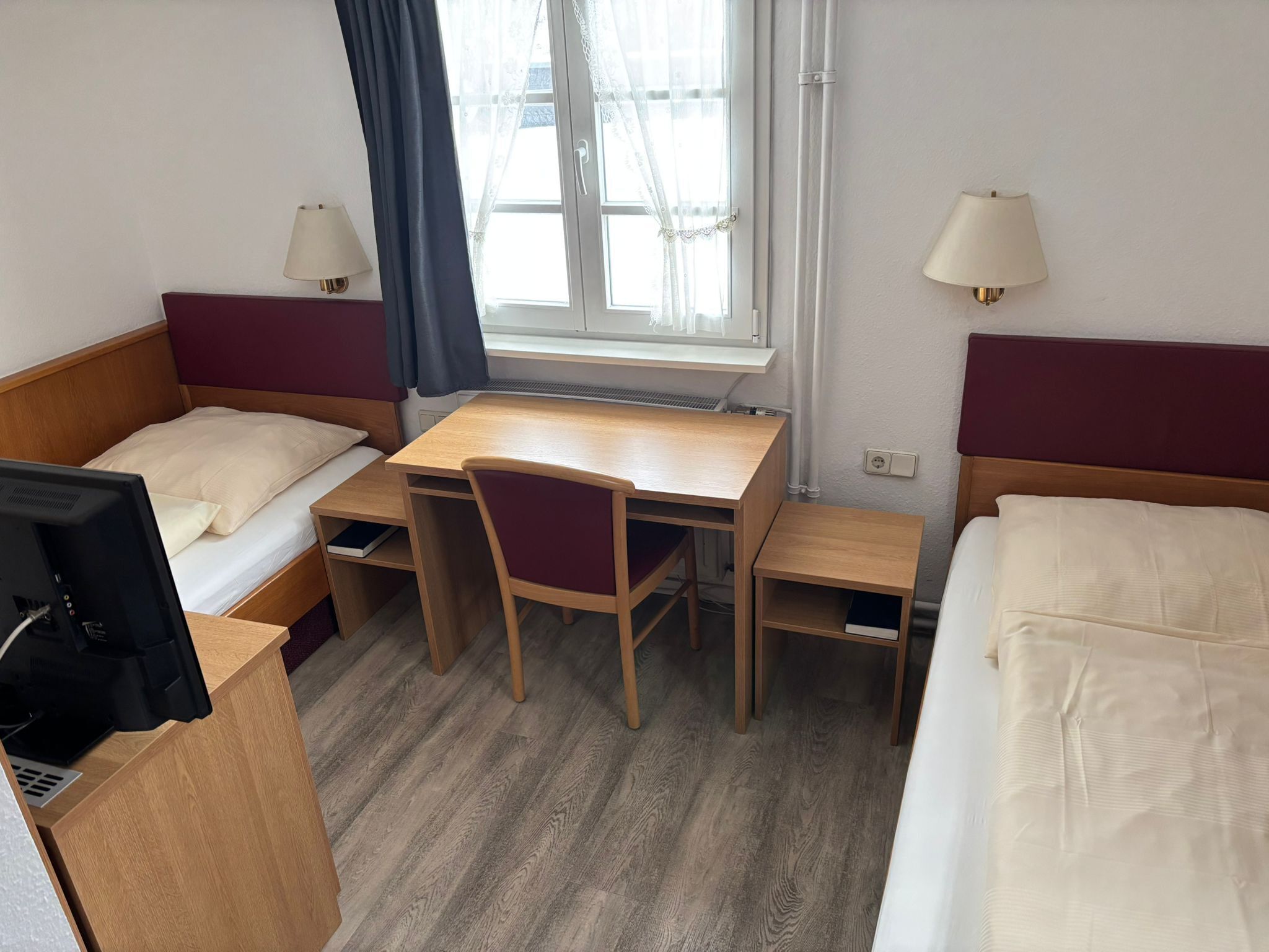 Habitación de hotel con dos camas individuales, un escritorio de madera, una silla, un televisor y dos lámparas de noche.