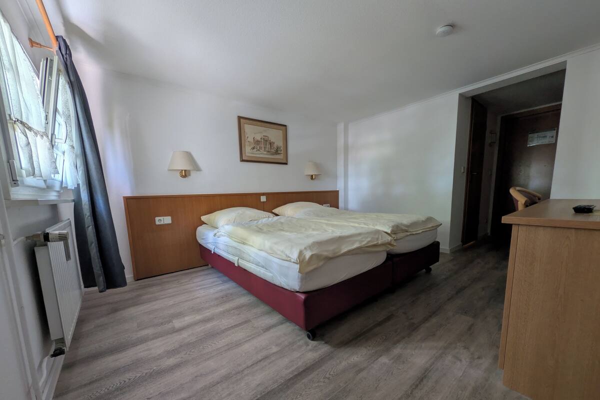 Une chambre d’hôtel avec un lit, une tête de lit en bois, un radiateur, un bureau et une porte.