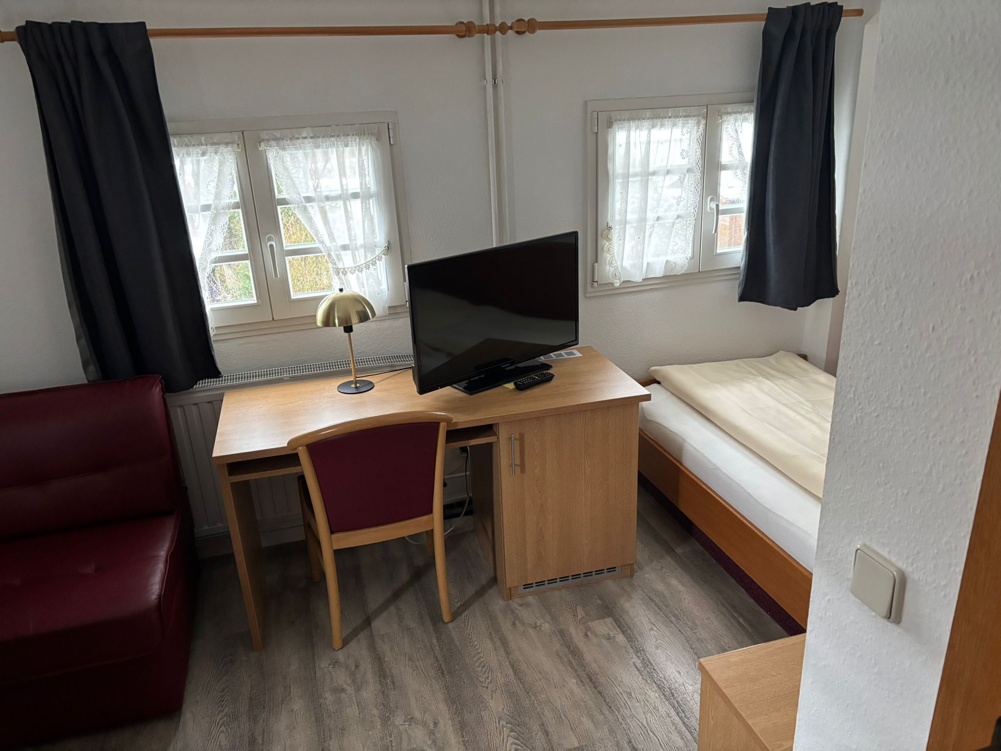 Una habitación compacta con un escritorio de madera, una cama individual, una silla roja y dos ventanas con cortinas.