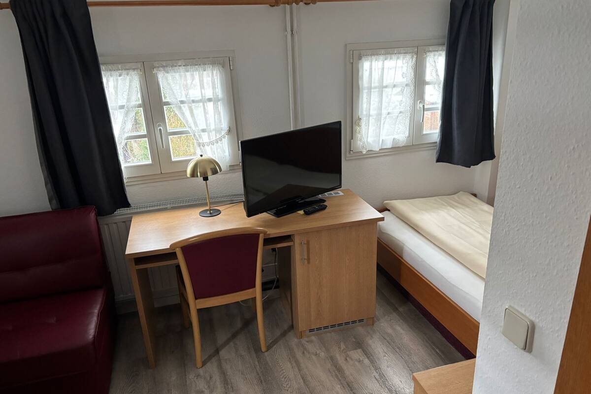 Une pièce de taille modeste, avec un bureau en bois, un lit simple, une chaise rouge et deux fenêtres équipées de rideaux.