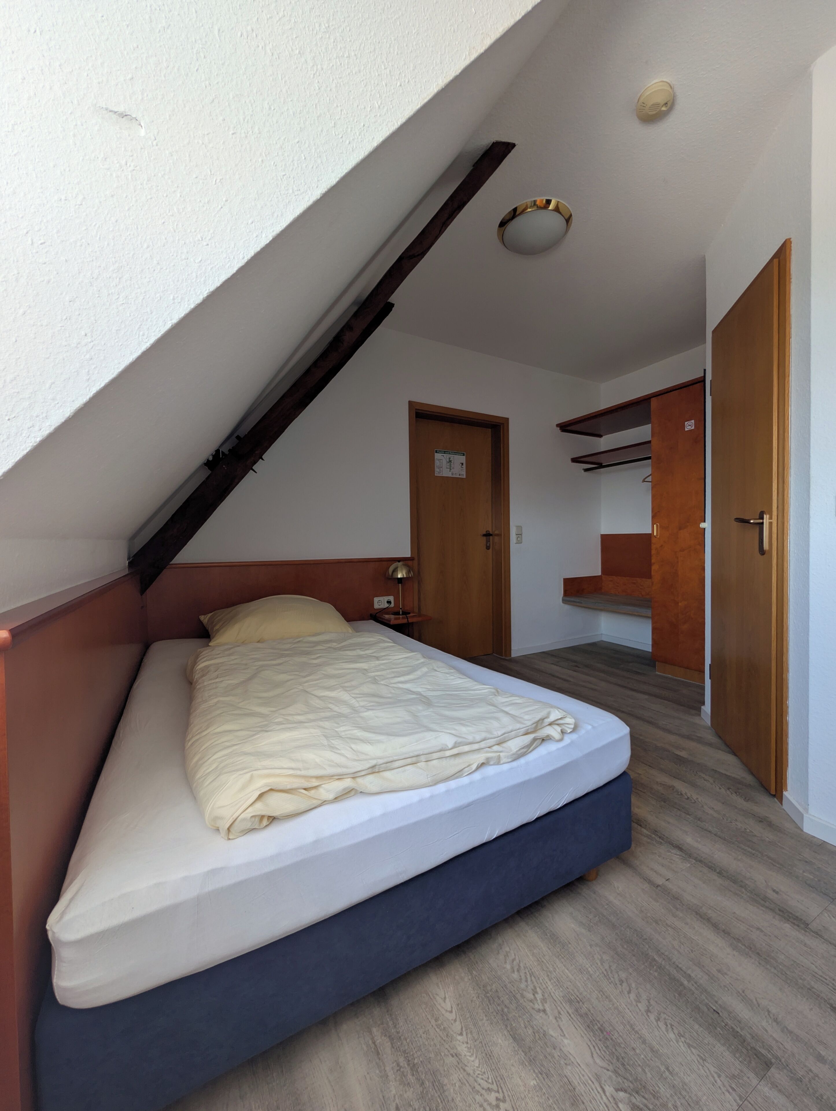 A bedroom with a sloped ceiling, a bed, a nightstand, a door, and a small shelf.