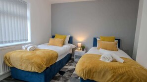 Ein Hotelzimmer mit zwei Betten, jeweils mit blauer Kopfteil, weißen Laken und einer gelben Decke. Auf jedem Bett befindet sich ein kleiner Nachttisch mit einer Lampe und einem zusammengeklappten Handtuch.