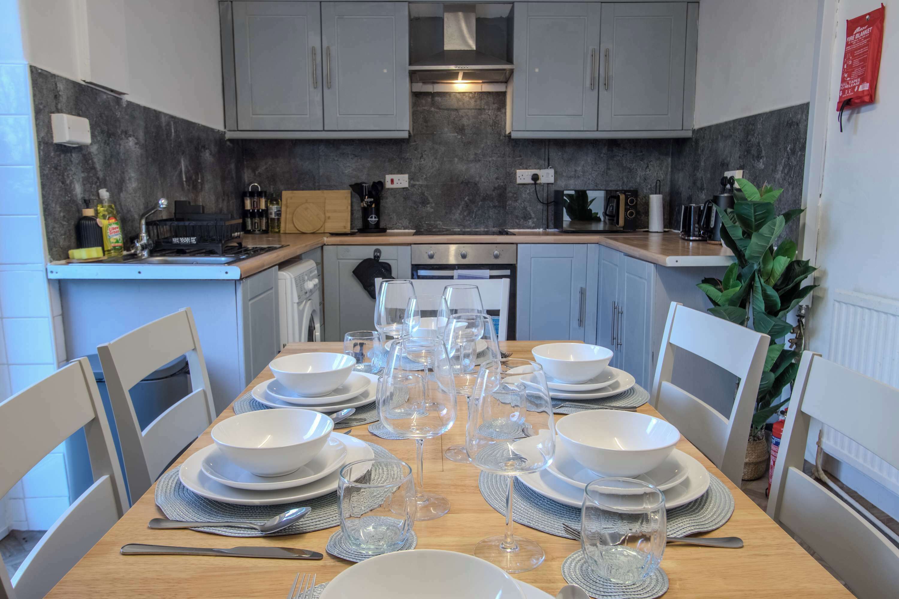 Une cuisine moderne avec des meubles gris, un plan de travail en bois et une table à manger dressée pour six personnes, avec de l’assiette blanche, des verres et des couverts.