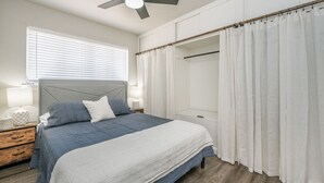 Un dormitorio con cama, mesitas de noche, ventilador de techo, ventana con persianas y vestidor con cortina.