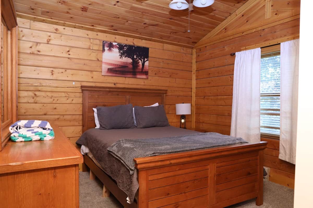 A wooden-paneled room with a bed, a nightstand, a lamp, and a window with curtains.