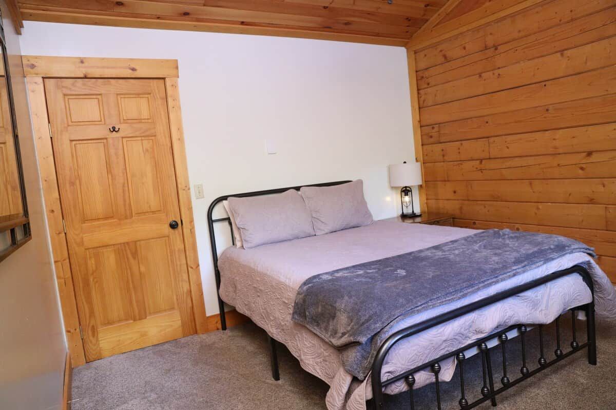 A bedroom with a wooden door, a bed with a metal frame, and a nightstand with a lamp.