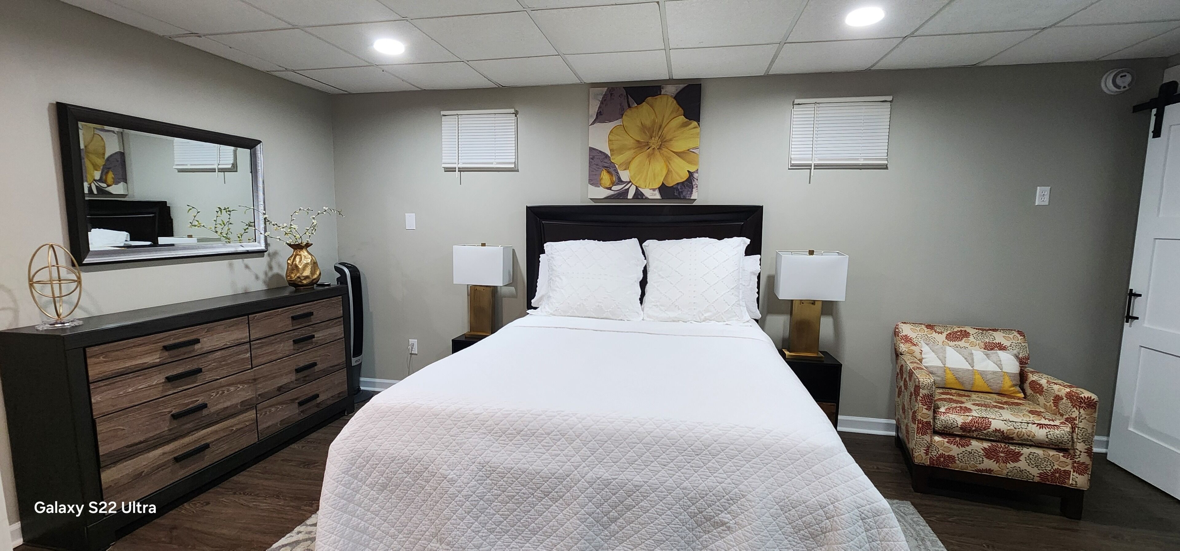 Une chambre à coucher avec un lit orné d’un motif inspiré du Galaxy S22 Ultra, une commode, deux lampes de chevet, un fauteuil et un grand tableau représentant des fleurs accroché au mur.