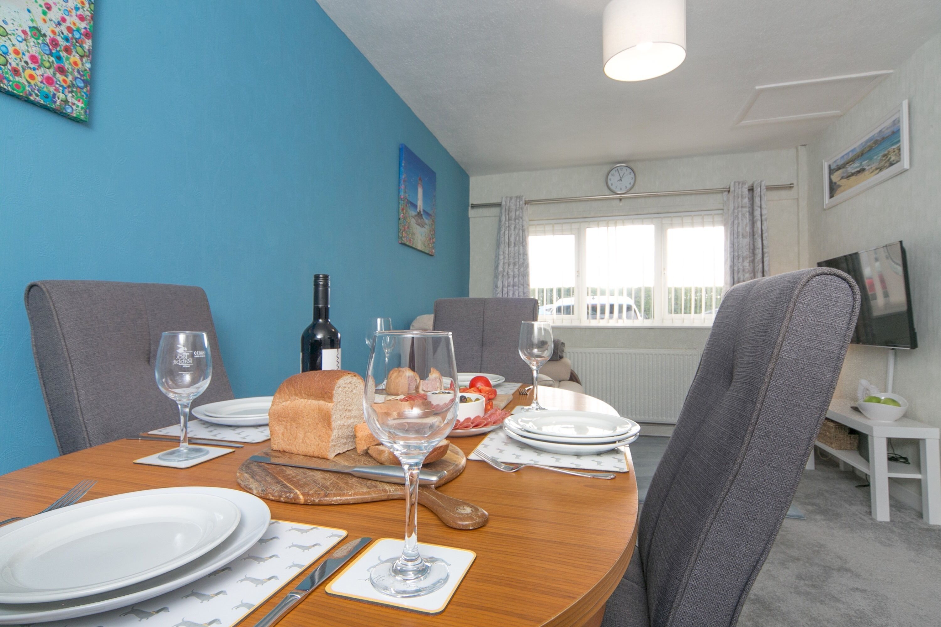 A dining area with a wooden table set for two, featuring plates, glasses, bread, and a bottle of wine.