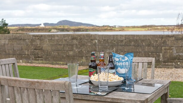 A table with a bowl of popcorn, two beer bottles, and a glass of beer, set against a backdrop of a stone wall and a body of water with mountains in the distance.