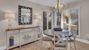 Een eetruimte met een ronde tafel, witte stoelen, een kroonluchter en een witte console tafel met een spiegel en lampen.