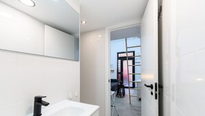 Une salle de bain moderne avec un lavabo blanc, un robinet noir et un miroir au-dessus. Une porte donne accès à un coin repas avec une table et des chaises.