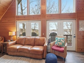 A cozy cabin interior with a leather sofa, wicker chair, and a large window overlooking a deck.