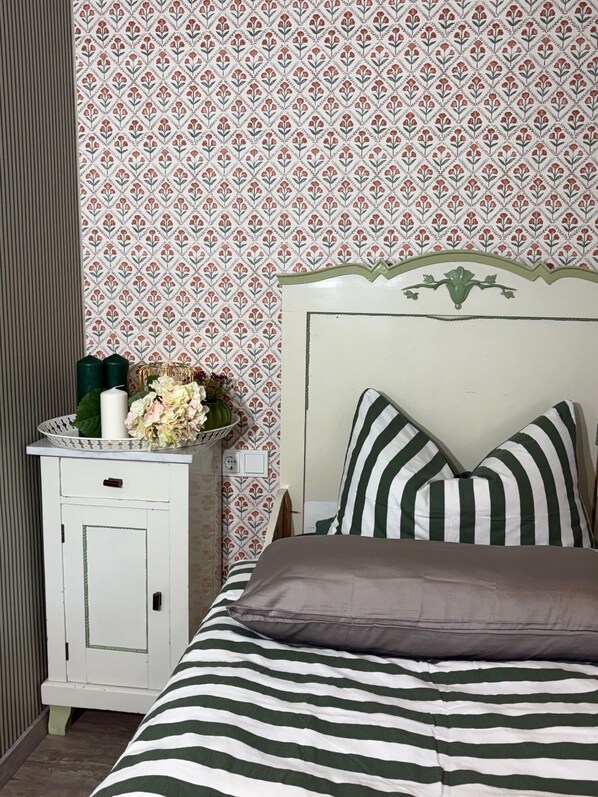 Quarto com colcha listrada, mesa de cabeceira branca com um vaso de flores e papel de parede estampado.