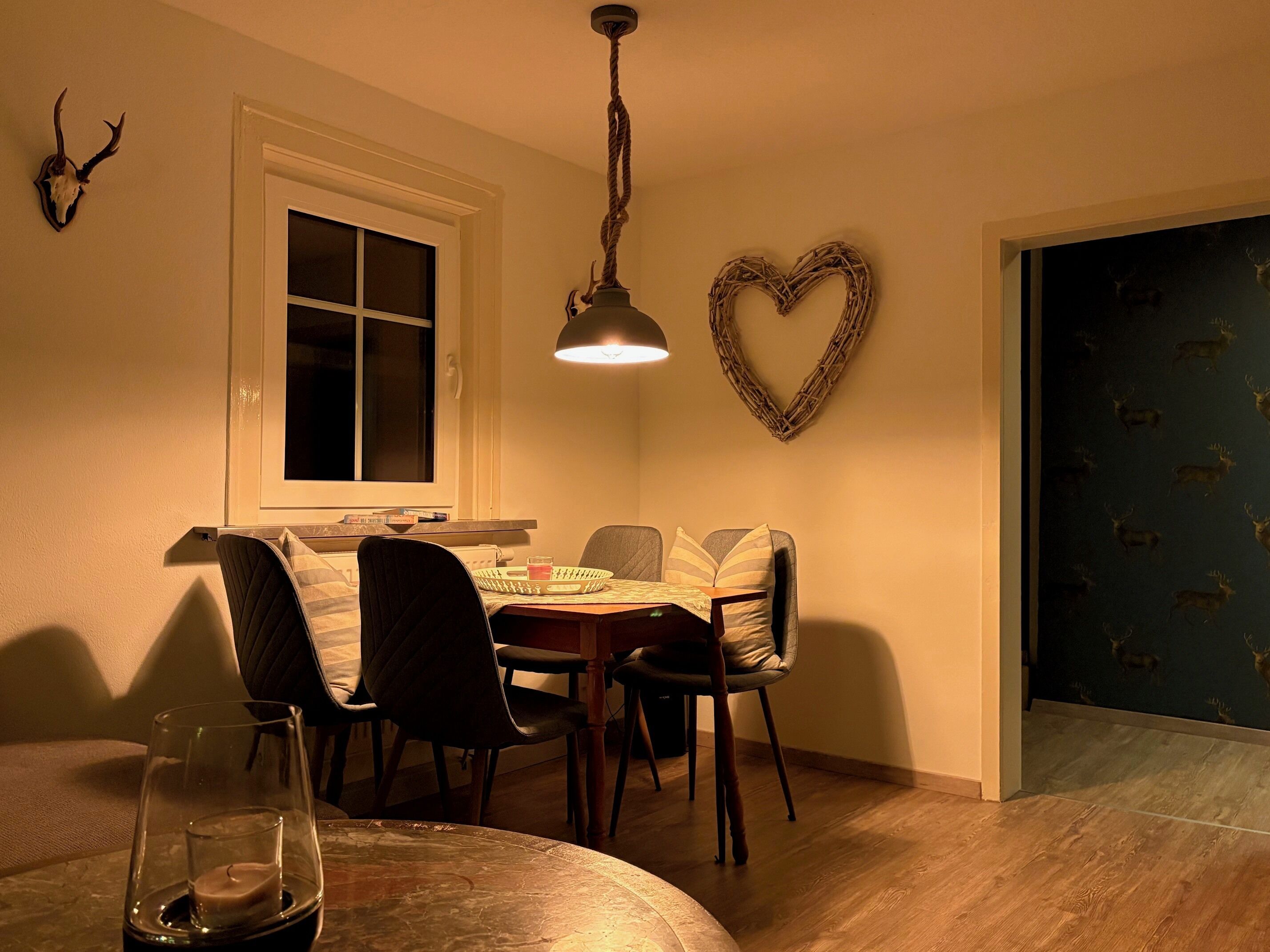 Un coin repas avec une table ronde, des chaises, un luminaire suspendu, une décoration en forme de cœur et une fenêtre avec des stores.