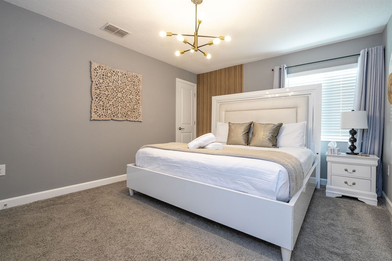 A modern bedroom with a white bed, a decorative wall piece, a ceiling light fixture, and a nightstand with a lamp.