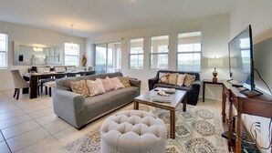 Ein geräumiges Wohnzimmer mit grauem Sofa, einem Couchtisch und einem Fernseher. Es gibt einen Essbereich mit Tisch und Stühlen sowie eine Küche im Hintergrund.