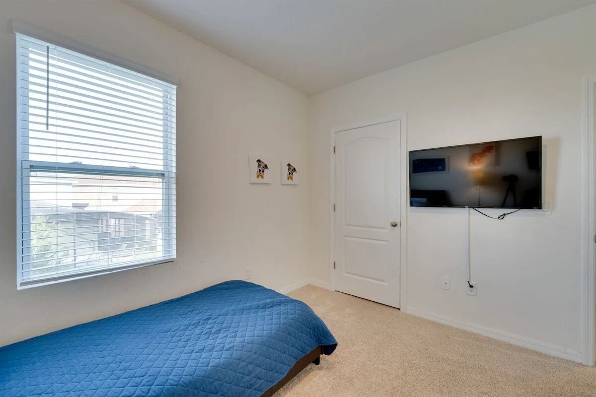 A bedroom with a single bed, a television mounted on the wall, a door, and a window with blinds.