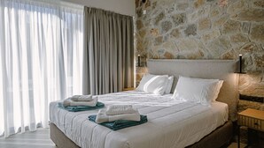 Moderni hotellihuone, jossa on kivestä tehty koristeellinen seinä, suuri sänky valkoisilla vuodevaatteilla ja näkymä ohuiden verhojen läpi.