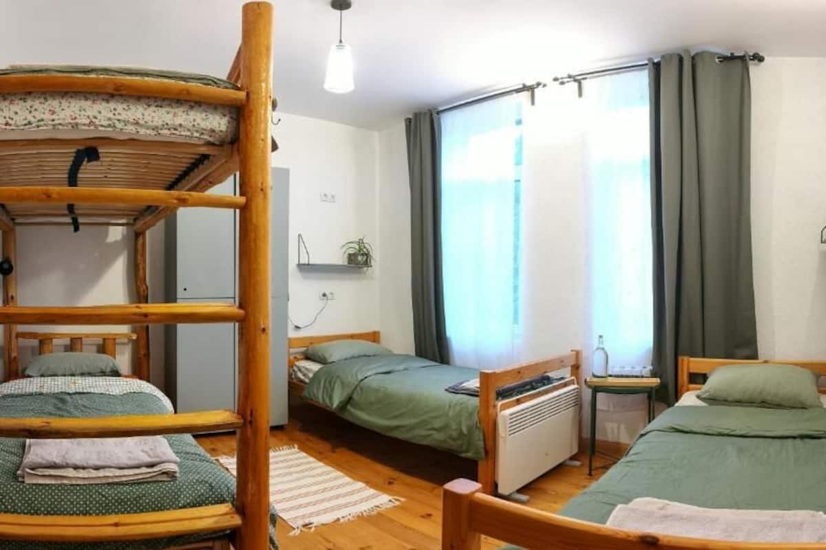 Una camera del dormitorio con letti a castello, un letto singolo, un termosifone, un tavolino e una finestra con tende.