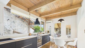 A modern kitchen with a stone wall, wooden ceiling, and a dining area with white chairs. - Charming country house near Lake Leman (Lully)