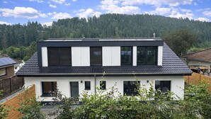 Une maison moderne à deux étages, dotée d’un toit en pente, de grandes fenêtres et offrant une vue sur une chaîne de montagnes boisée.